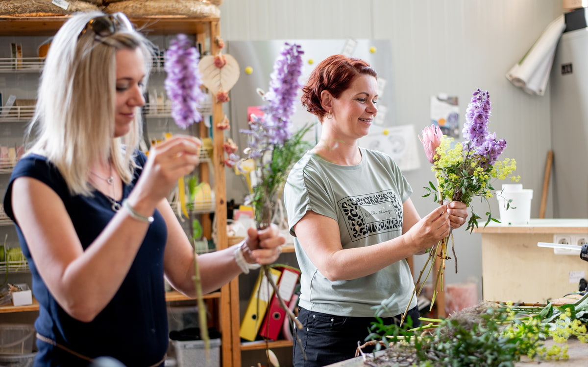Zwei Floristinnen arrangieGartenarbeiter arrangiert Pflanzen und Produkte auf Verkaufstischen im Gartencenterren frische Blumen in einer Werkstatt; im Vordergrund bindet eine Frau ein Bouquet, während im Hintergrund weitere Blumen vorbereitet werden. Zwei Floristinnen arrangieGartenarbeiter arrangiert Pflanzen und Produkte auf Verkaufstischen im Gartencenterren frische Blumen in einer Werkstatt; im Vordergrund bindet eine Frau ein Bouquet, während im Hintergrund weitere Blumen vorbereitet werden.